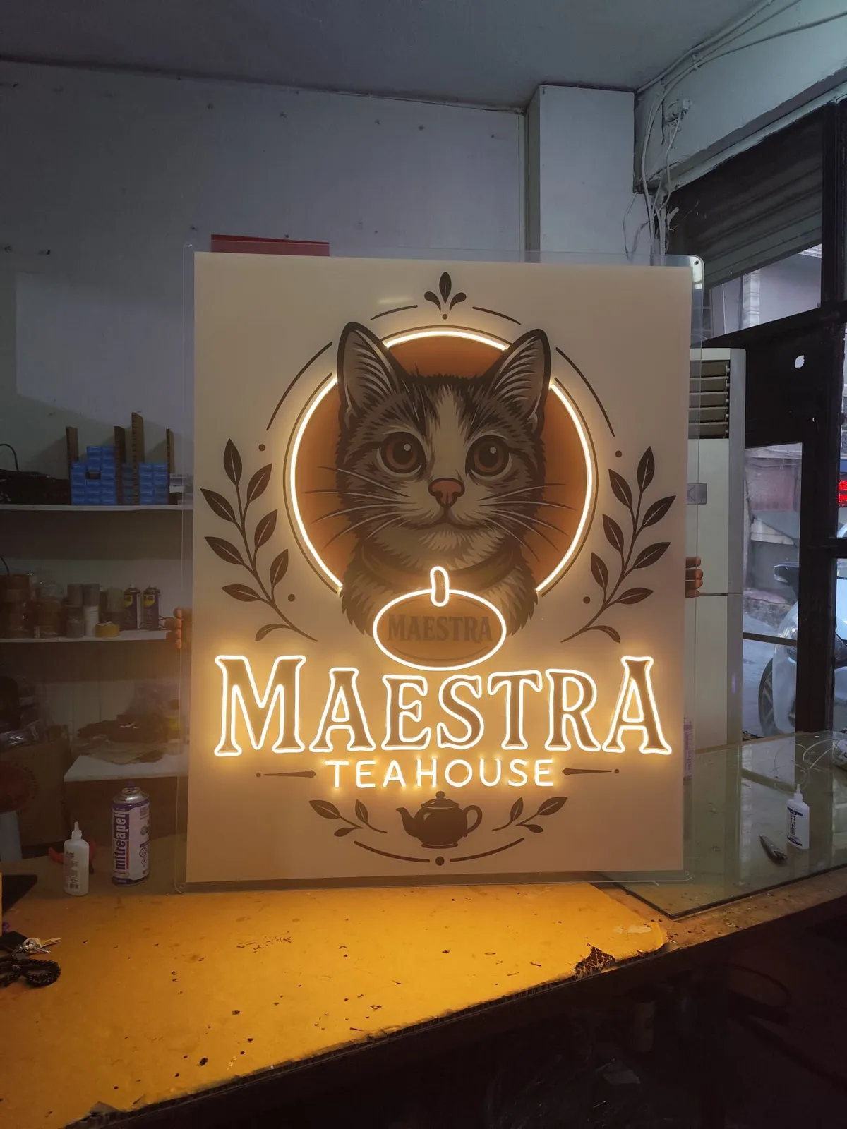 Warm white 'Maestra Teahouse' logo neon on a printed cat illustration backboard